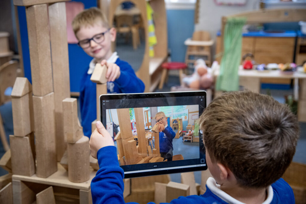 child holding an ipad and filming another who is building with blocks