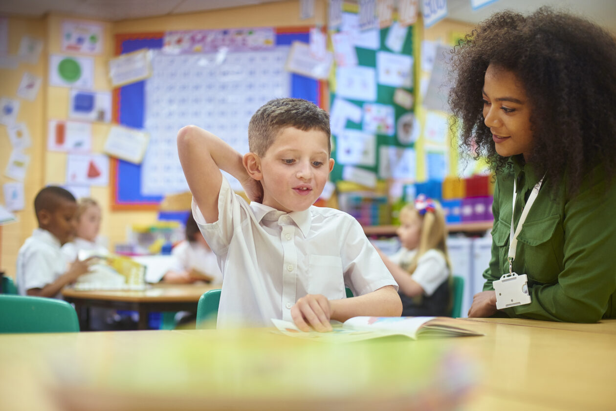 a primary aged schoolboy sits with his teacher in classroom setting. both are smiling. other children are visible in the background of the image, sitting round a classroom table doing some work.