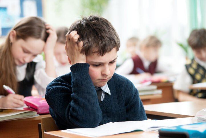 A school aged boy sits in a crowded classroom at a single desk. He has one arm on the desk, propping up his head and a difficult expression on his face.