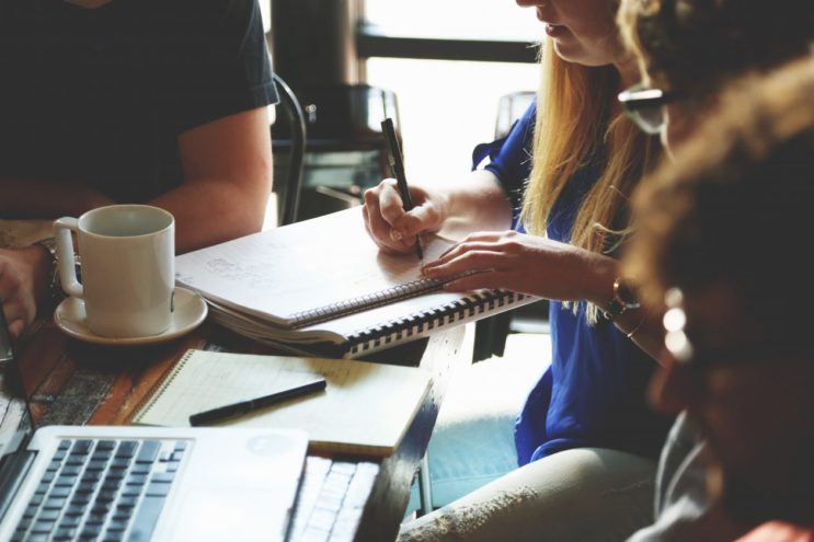 Close up image of female writing notes in a notebook alongside colleagues seated at a table