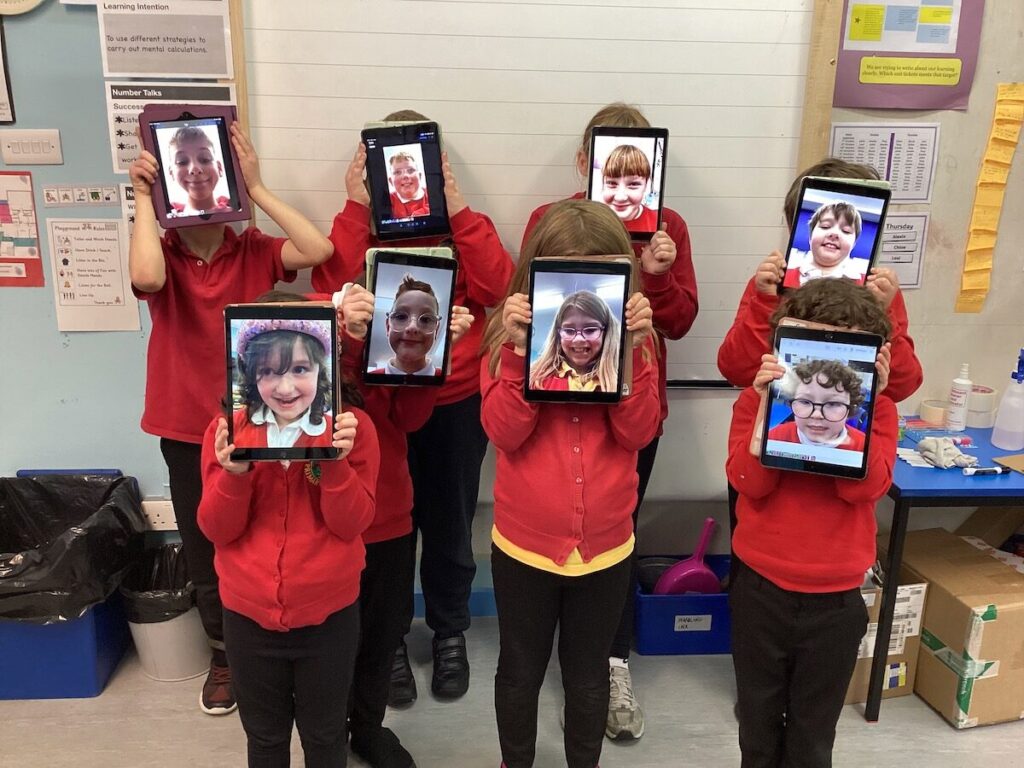 A group of children holding up iPads in front of their faces. The iPad scheens show children's photogtaphs.