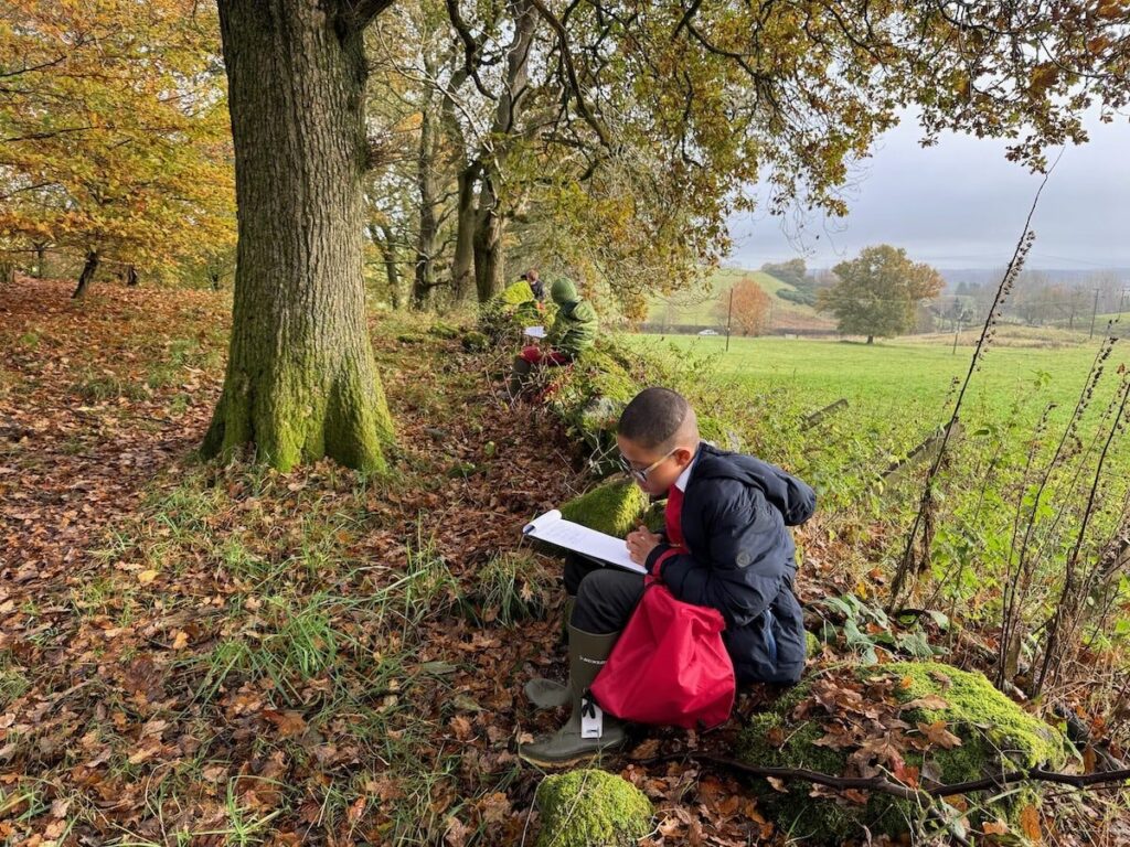 Children sitting along  at the edge of a wood a wall writing.