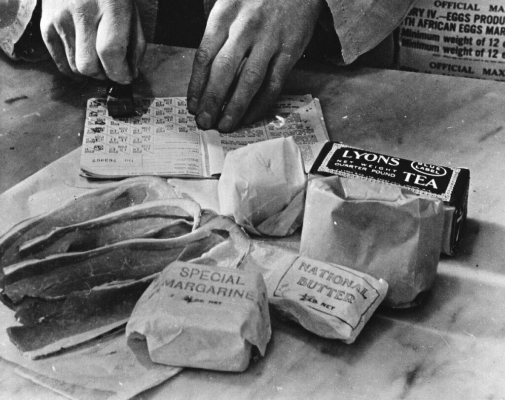 Posed photograph in which a shopkeeper cancels the coupons in a British housewife's ration book for the tea, sugar, cooking fats and bacon she is allowed for one week. Most foods in Britain are rationed and some brand names are given the designation "National".